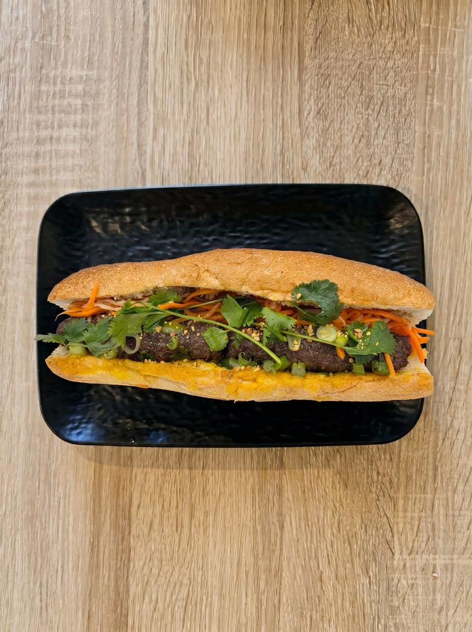 A Vietnamese banh mi sandwich with grilled pork, cilantro, carrots, and green onions on a black plate.