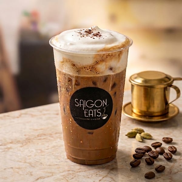 Iced Vietnamese coffee with foam and Saigon Eats logo, next to coffee beans and a phin filter.