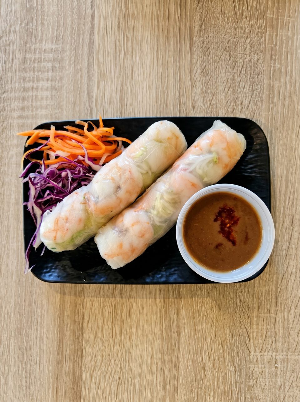 Two shrimp spring rolls with shredded carrots, purple cabbage, and peanut dipping sauce on a black plate.