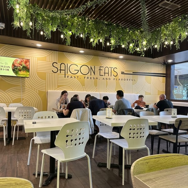 Diners seated at tables inside Saigon Eats, with a yellow patterned wall and hanging greenery.