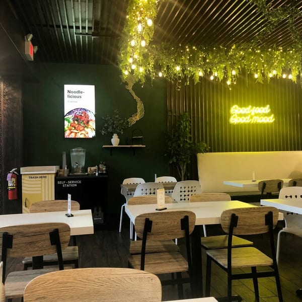 Modern restaurant interior with dark green walls, light wood tables and chairs, and glowing neon signs.