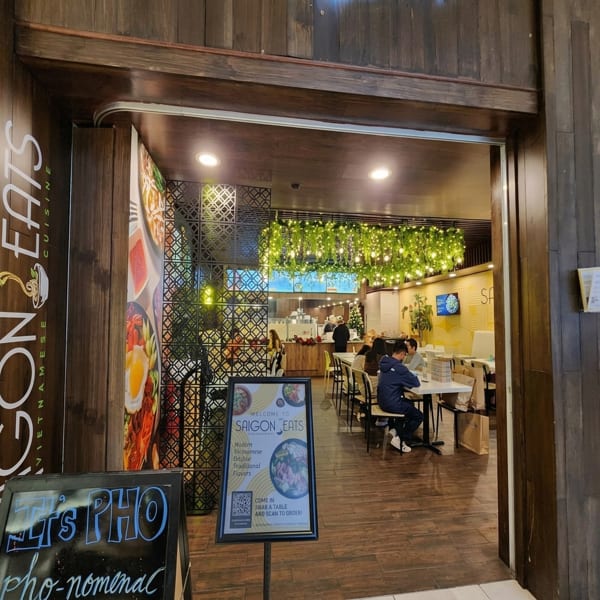Entrance to Saigon Eats, a Vietnamese restaurant, with customers dining inside under a leafy light fixture.